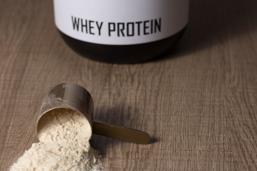 Whey protein food supplement for training and exercise. Dropped scoop with vanilla powder flavour. Wooden table. Black jar behind