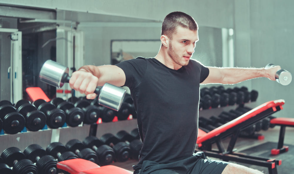 Athletic young man exercising his shoulders and doing side raises with pair of dumbbells at gym