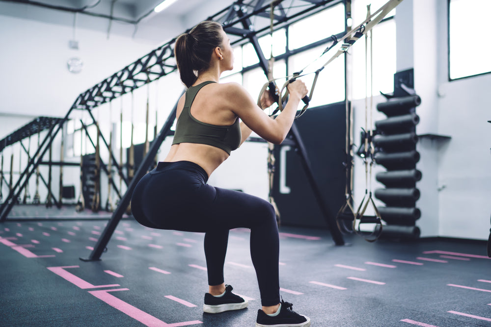 Side view of young fitness woman in sport top and leggings squatting with TRX straps during functional training in gym