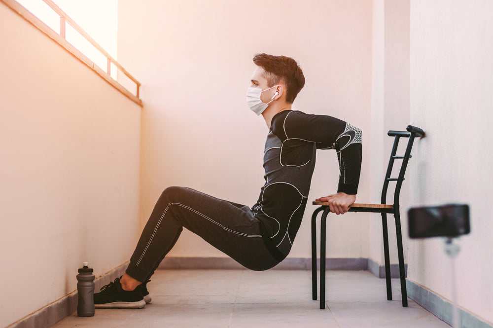 Young sports man in protective mask doing triceps dips on chair 