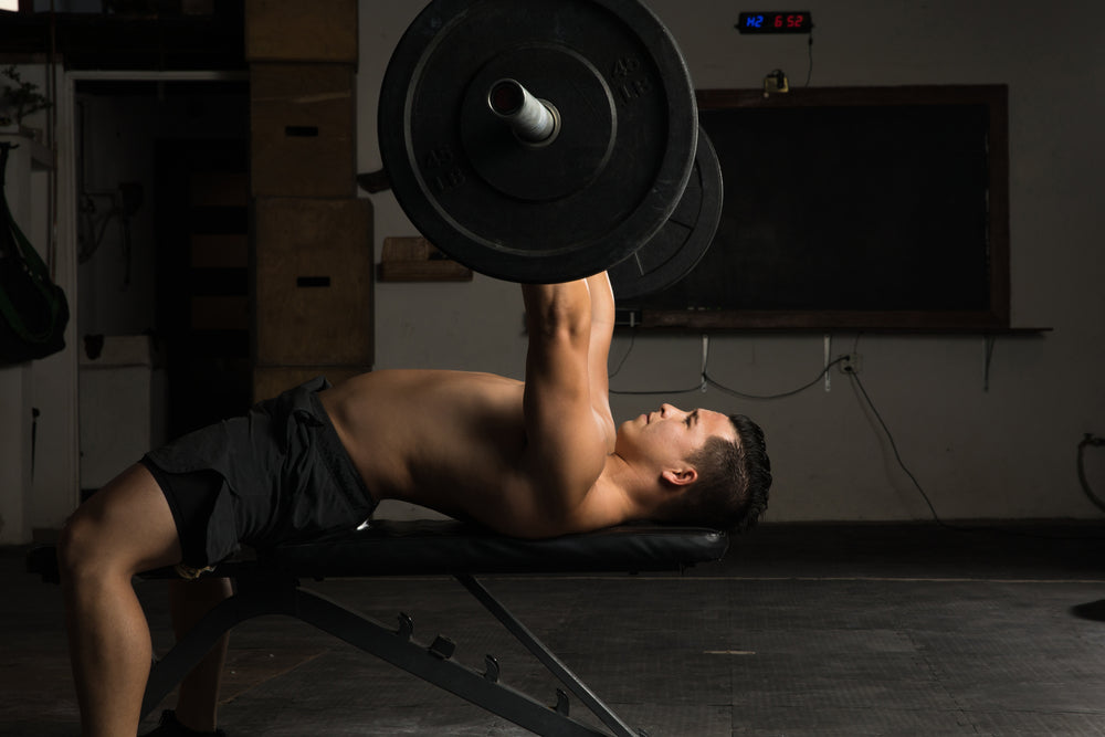 Profile view of a strong shirtless man doing bench presses in a cross-training gym