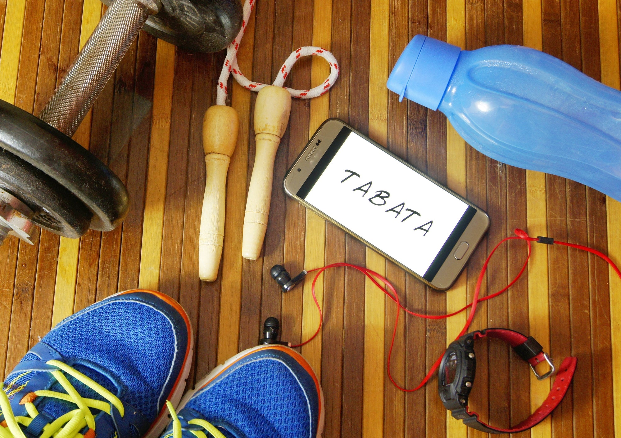 Dumbbells, sport shoes, smart phone with earphone, skipping rope and water bottle on wooden gym floor with word tabata