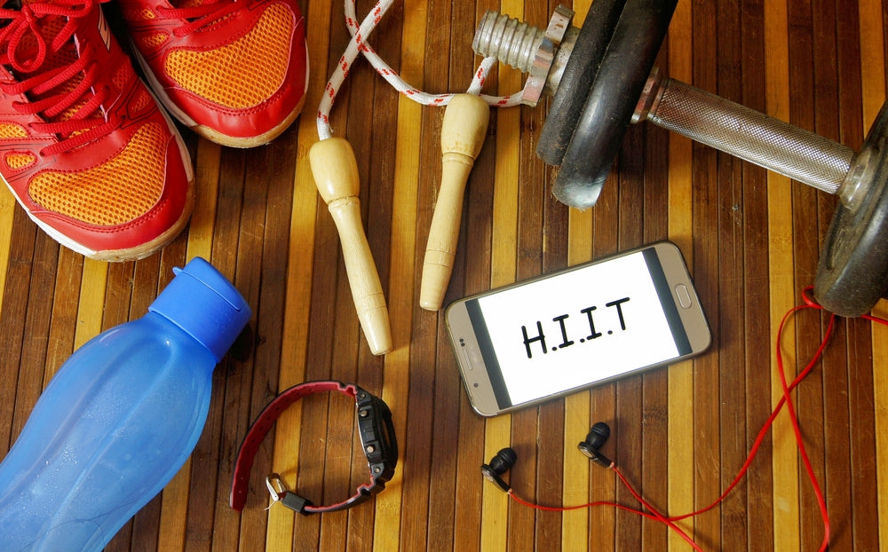 Dumbbells, sport shoes, smart phone with earphone, skipping rope and water bottle on wooden gym floor with word HIIT